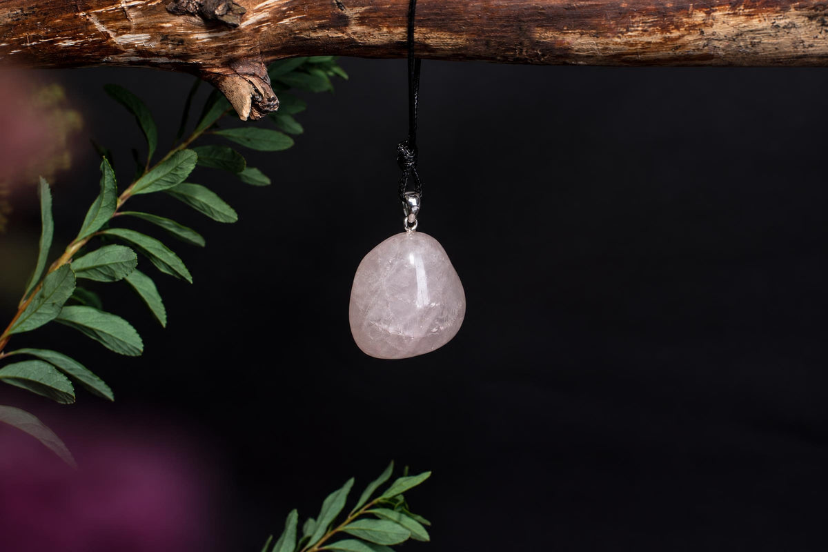 A pale pink, smooth stone pendant hangs from a black cord on a dark surface, with green leaves nearby.