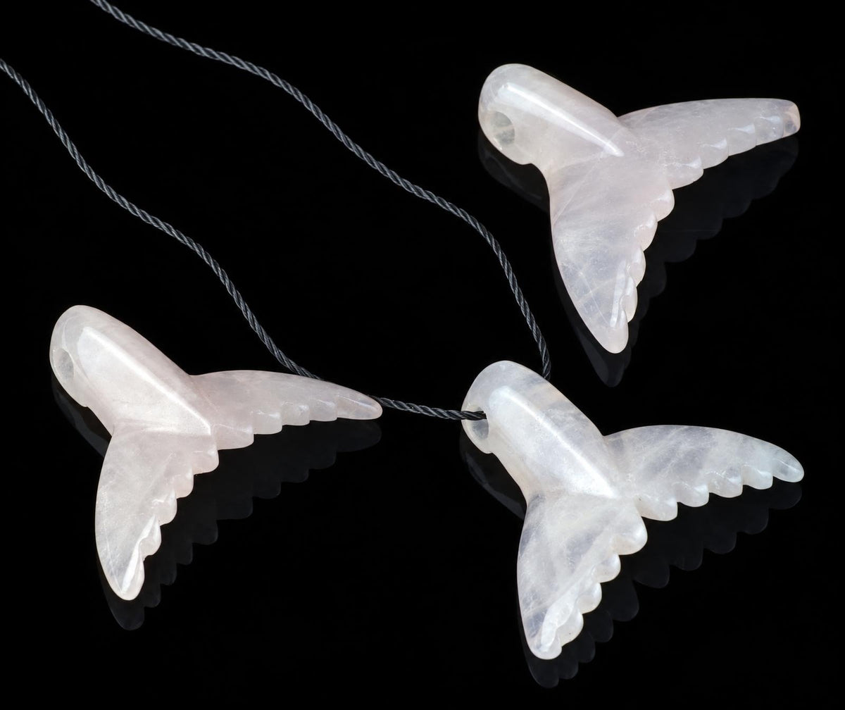 Three translucent, pale pink carved pendants shaped like dolphin tails hang from a dark cord on a black reflective surface.
