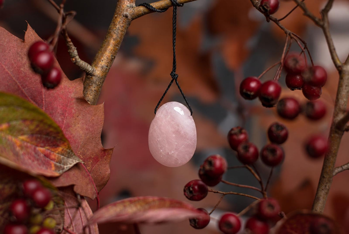 A smooth, pale pink oval stone pendant hangs from a black cord against a dark surface with red berries and autumn leaves.