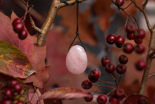 A smooth, pale pink oval stone pendant hangs from a black cord against a dark surface with red berries and autumn leaves.