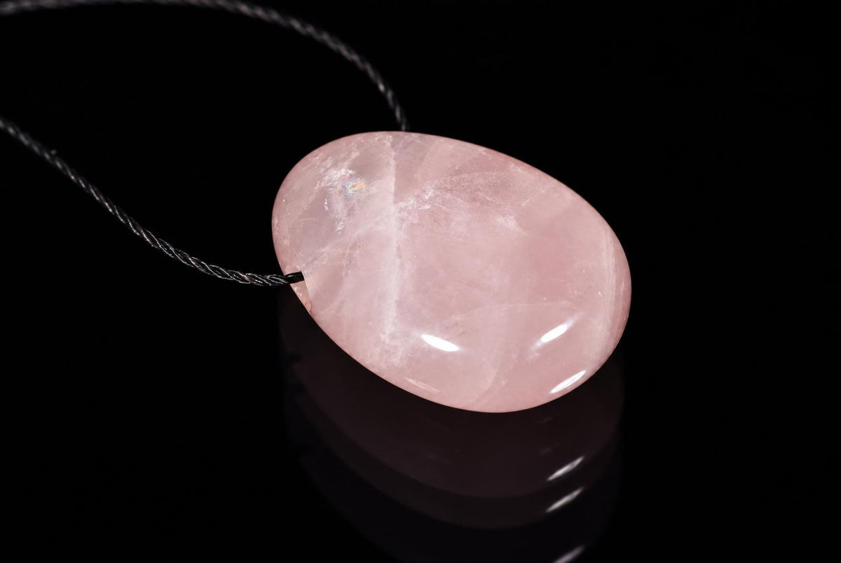 A smooth, oval-shaped pink stone pendant on a dark cord rests on a reflective black surface.