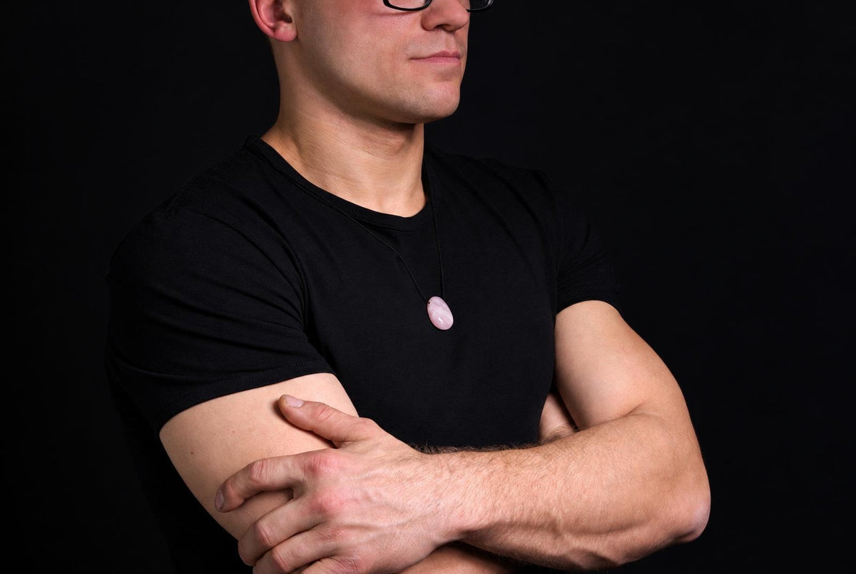 A man wears a black t-shirt and glasses, with arms crossed, displaying a smooth pink oval pendant on a dark surface.