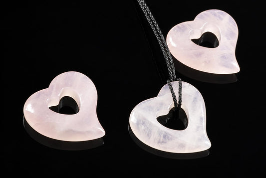 Three heart-shaped rose quartz pendants with a central hole, one on a dark cord, on a black surface.