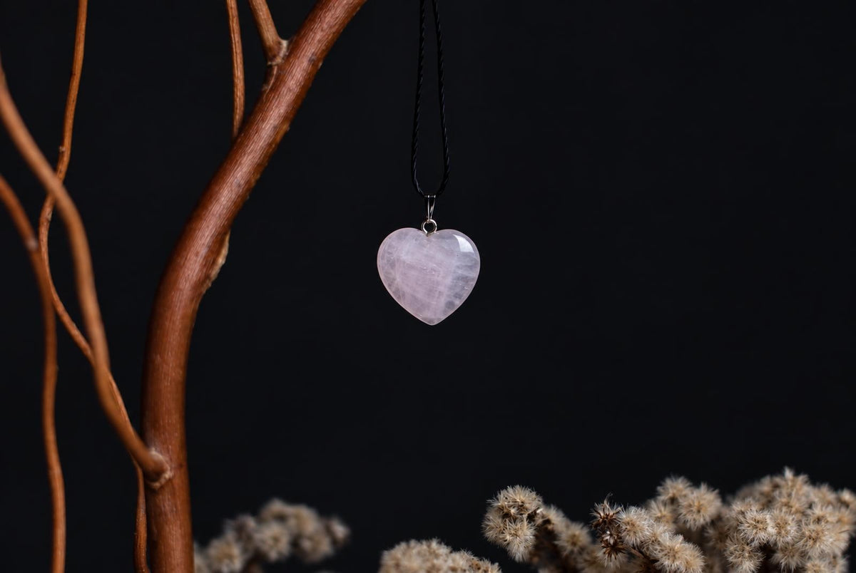 A heart-shaped rose quartz pendant hangs from a black cord against a dark background, with dried branches and flowers nearby.