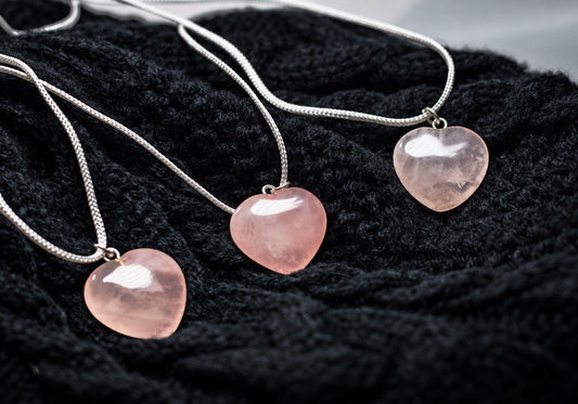 Three silver-toned necklaces with pink heart-shaped rose quartz pendants on a dark knitted surface.