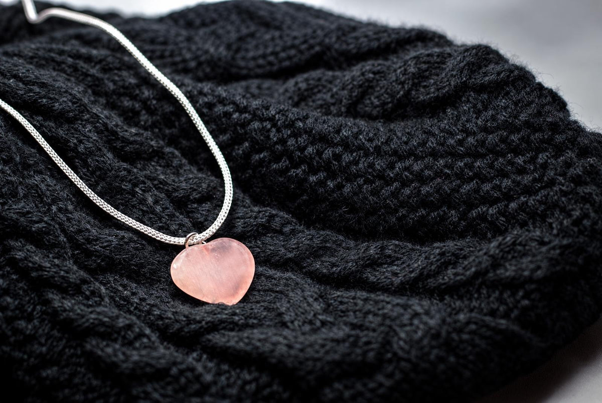 A silver-toned necklace with a pink heart-shaped pendant rests on a dark knitted fabric surface.