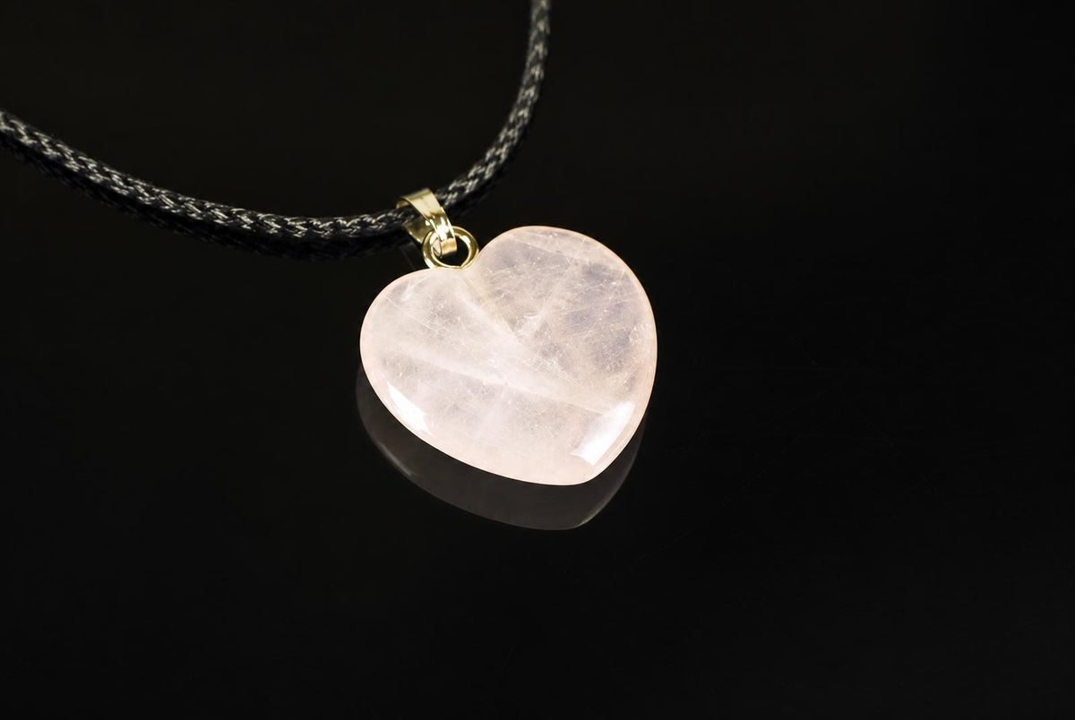 A heart-shaped rose quartz pendant with a gold-coloured bail hangs from a black and white braided cord against a dark surface.