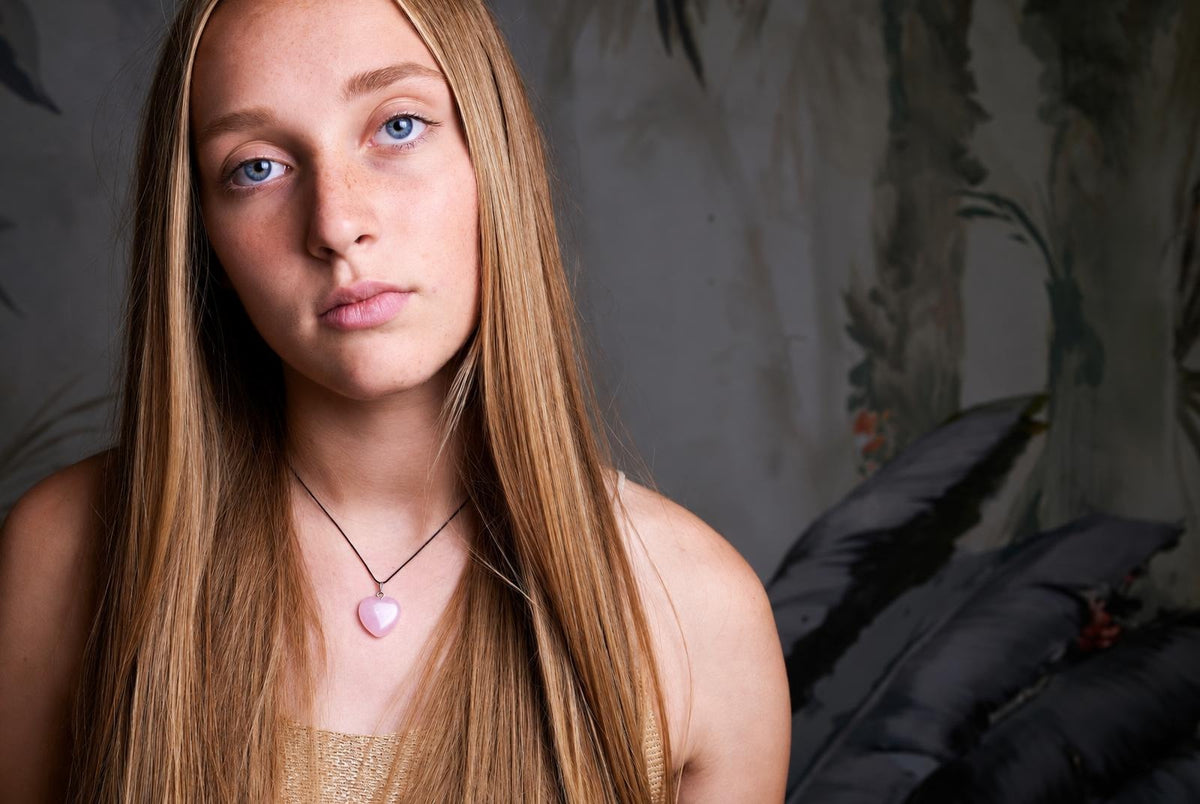 A person wears a necklace with a pink heart-shaped pendant on a dark cord against a patterned wall.