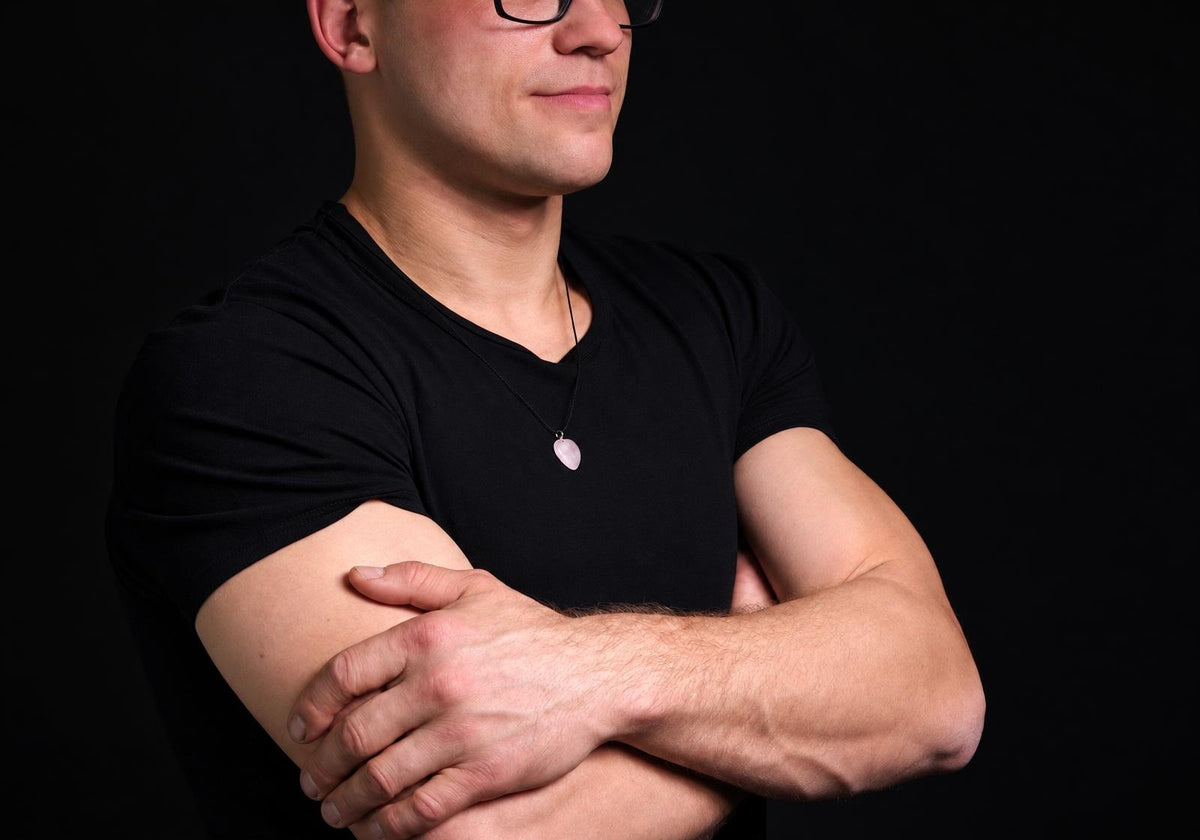 A man in a black t-shirt wears a necklace with a pale pink, heart-shaped pendant against a dark background.