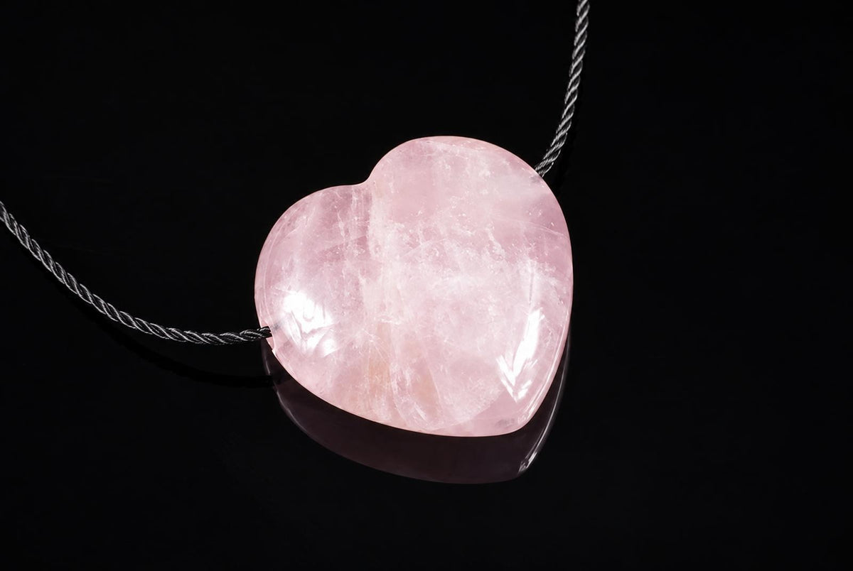 A polished pink heart-shaped pendant on a twisted silver-toned cord, resting on a dark reflective surface.