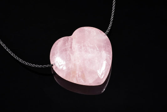 A polished pink heart-shaped pendant on a twisted silver-toned cord, resting on a dark reflective surface.
