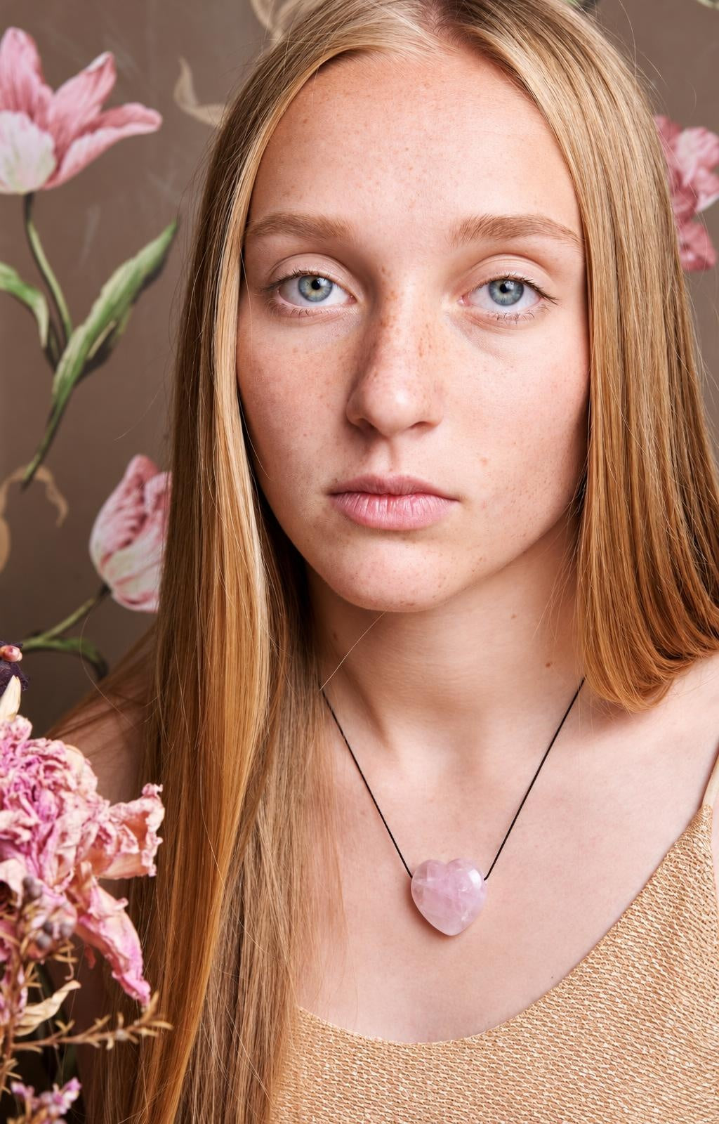 A woman wears a necklace with a pink, heart-shaped pendant on a black cord against a floral background.