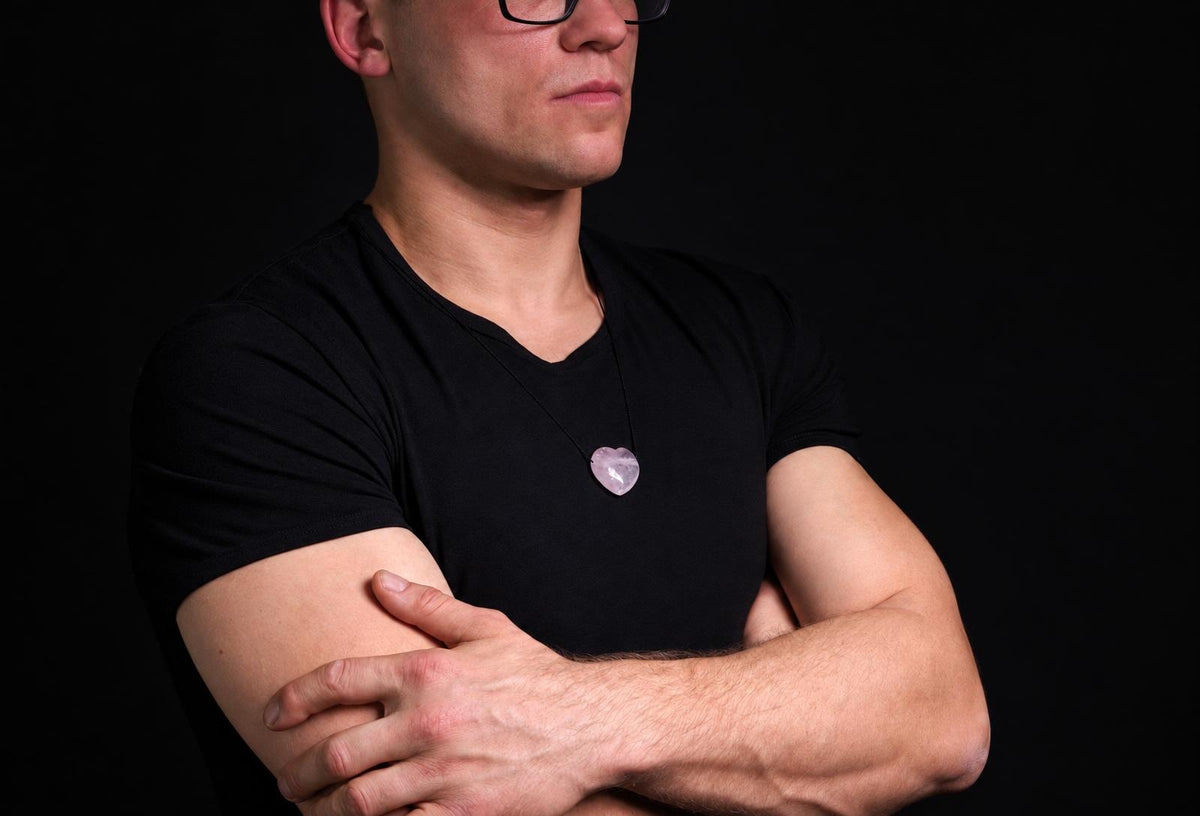 A man wears a black t-shirt with a heart-shaped, pale pink pendant on a dark surface.
