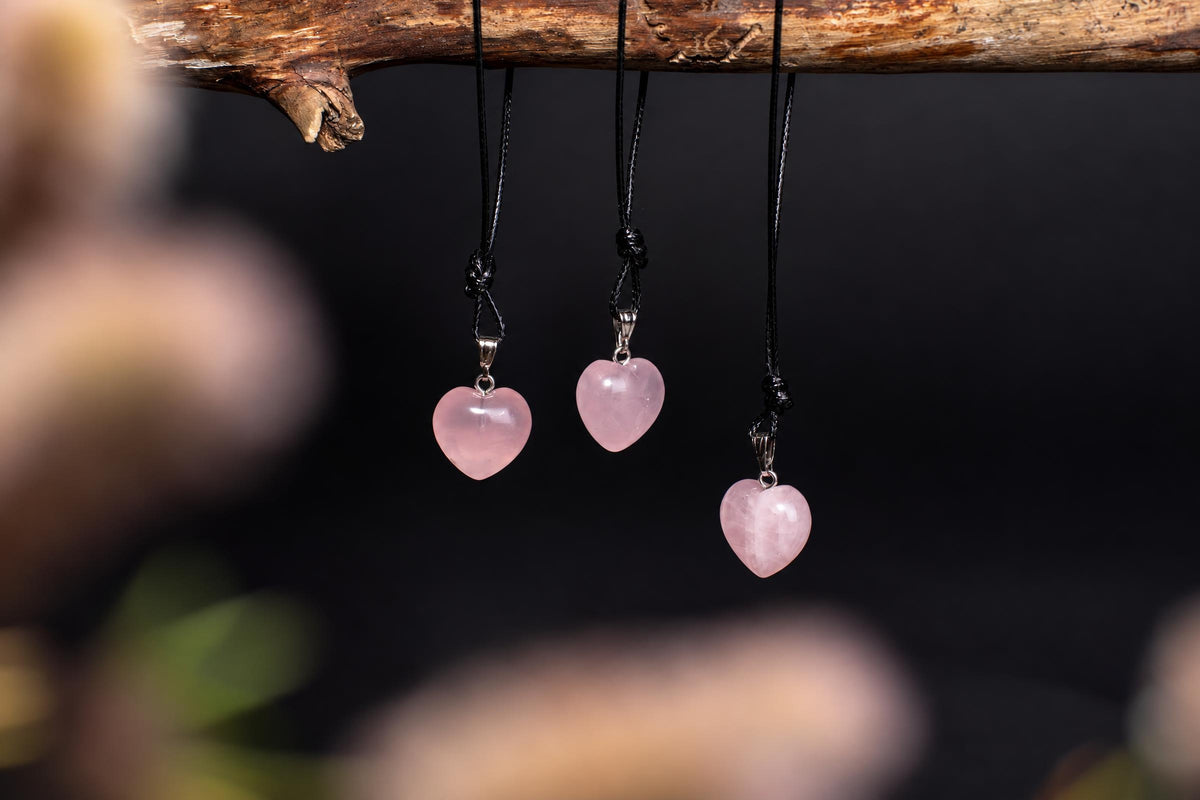 Three pink heart-shaped pendants hang from black cords on a wooden branch against a dark background.
