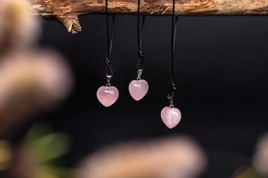 Three pink heart-shaped pendants hang from black cords on a wooden branch against a dark background.
