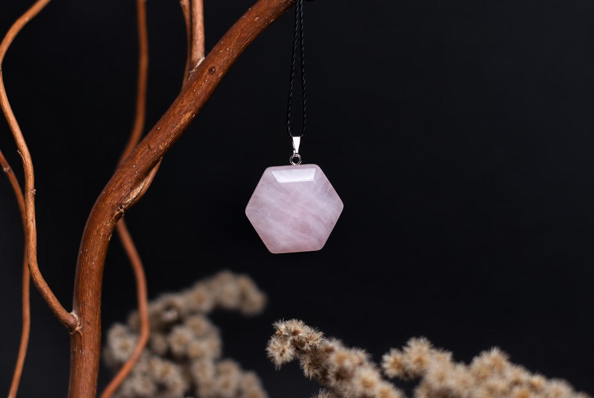 A hexagonal rose quartz pendant hangs from a black cord against a dark background, with dried branches and flowers nearby.
