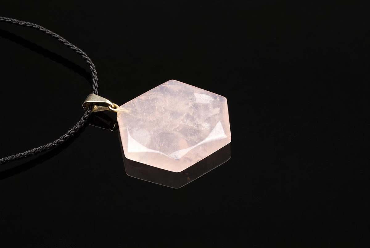 A pale pink rose quartz pendant has a faceted hexagonal shape and hangs from a black cord over a dark surface.
