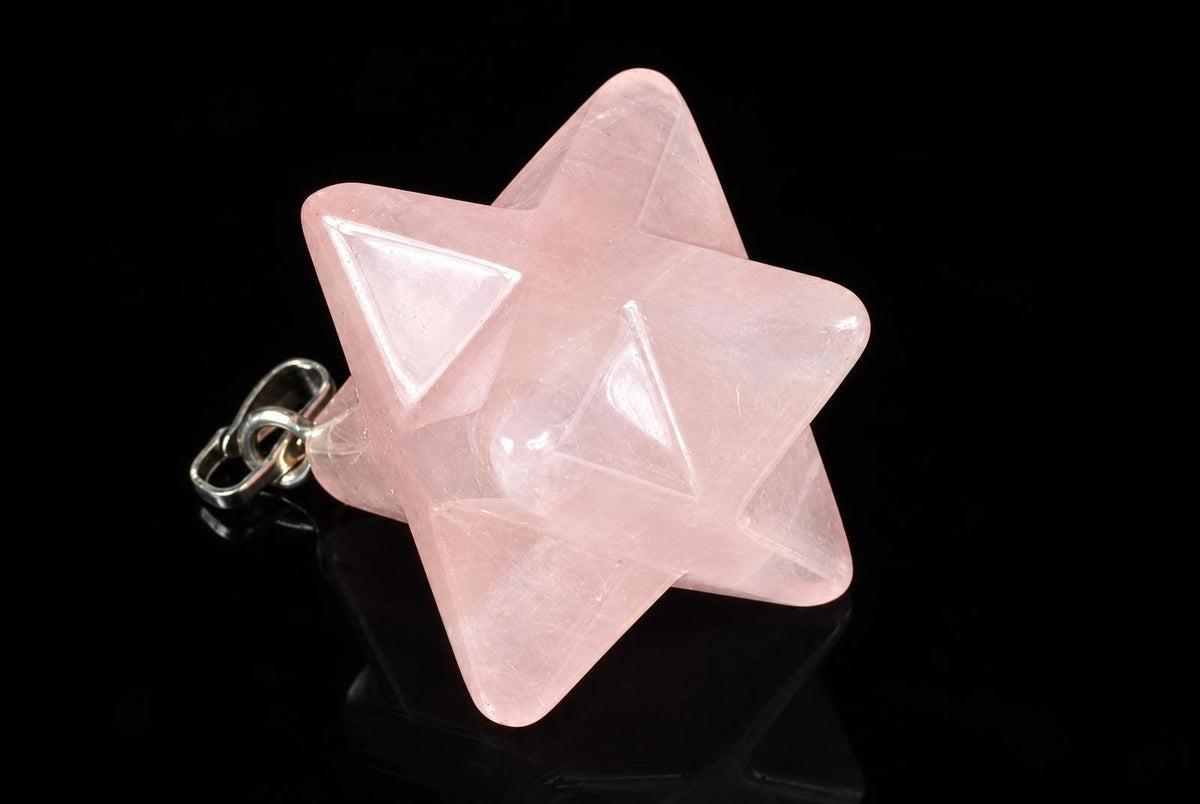 A pale pink rose quartz pendant forms a pointed geometric star with triangular faces and a metal loop on a glossy black surface.