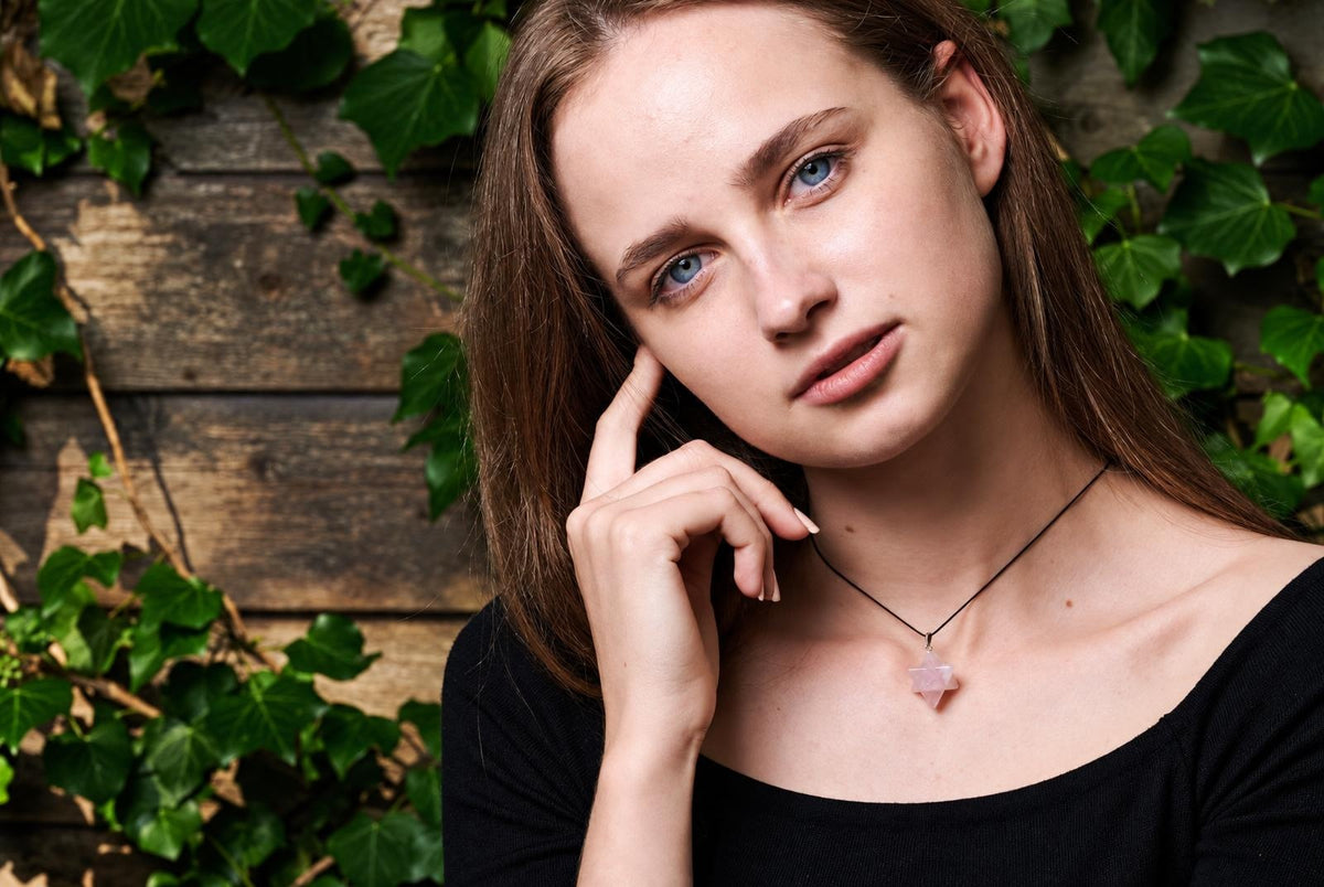 A woman wears a necklace with a pink, star-shaped pendant on a dark cord against a wooden background with green leaves.