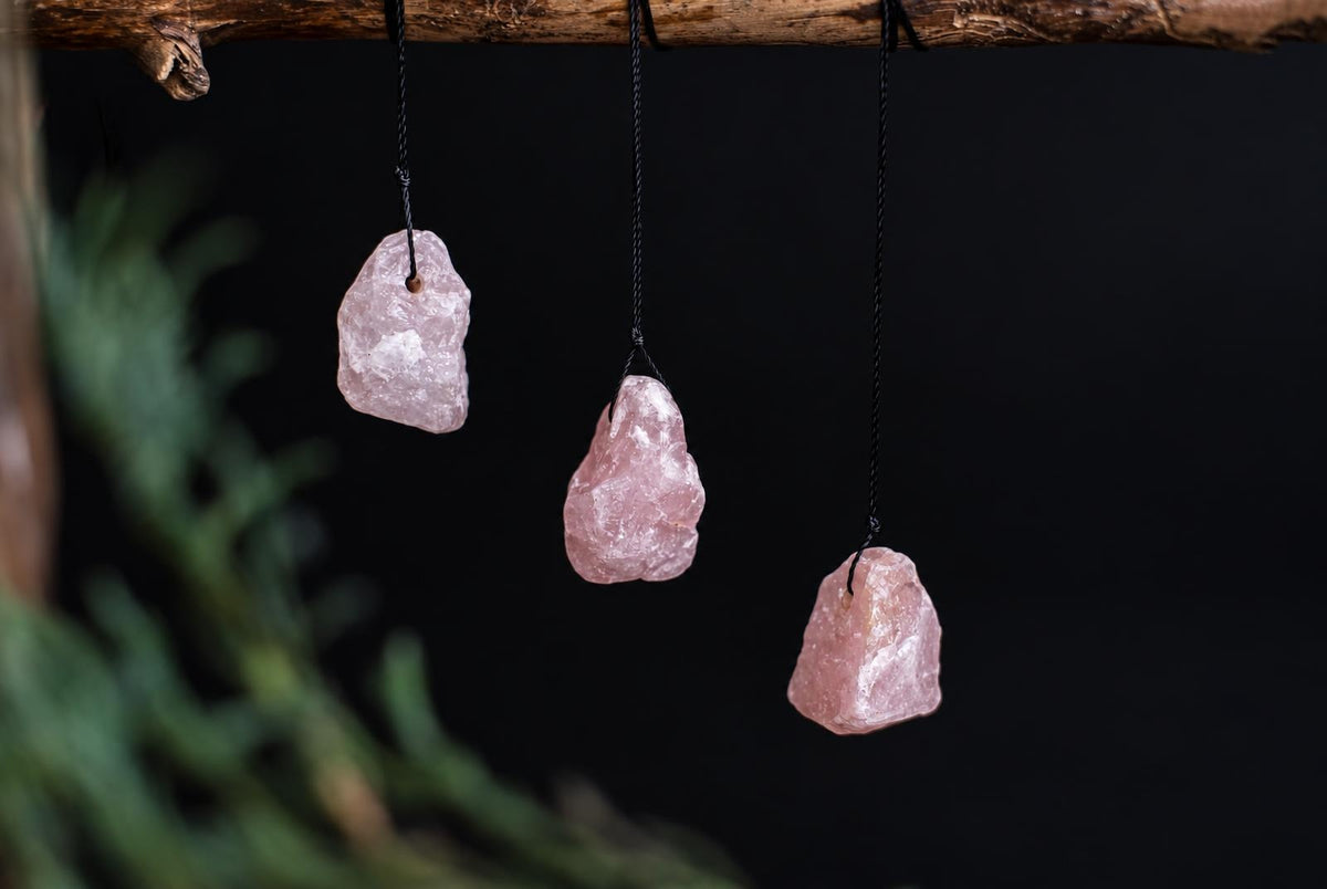 Three pink rose quartz stones hang from black cords on a wooden branch against a dark background.