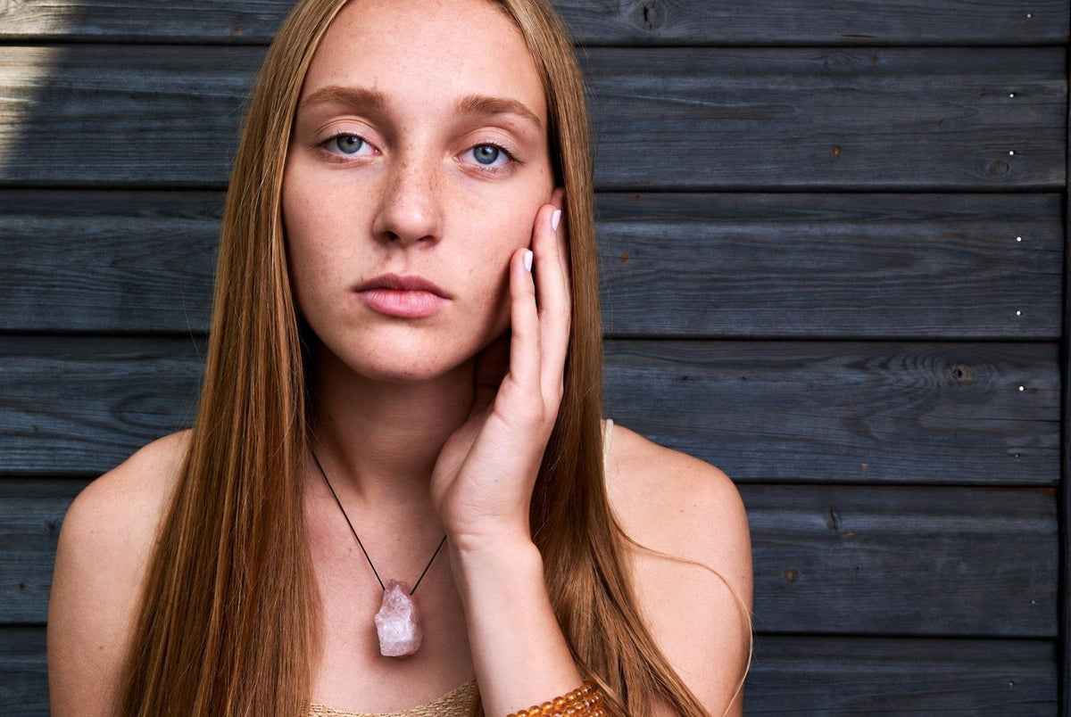 A woman with long hair wears a necklace with a pale pink, rough crystal pendant on a dark cord against a dark wooden background.