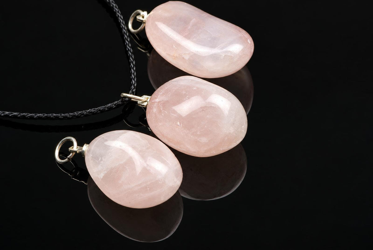 Three smooth, pale pink pebble-shaped pendants on dark cords rest on a black reflective surface.