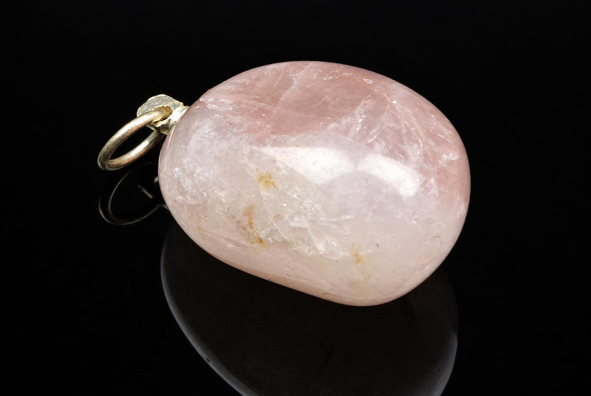 A polished pink rose quartz pebble pendant with a gold-coloured bail, resting on a dark reflective surface.