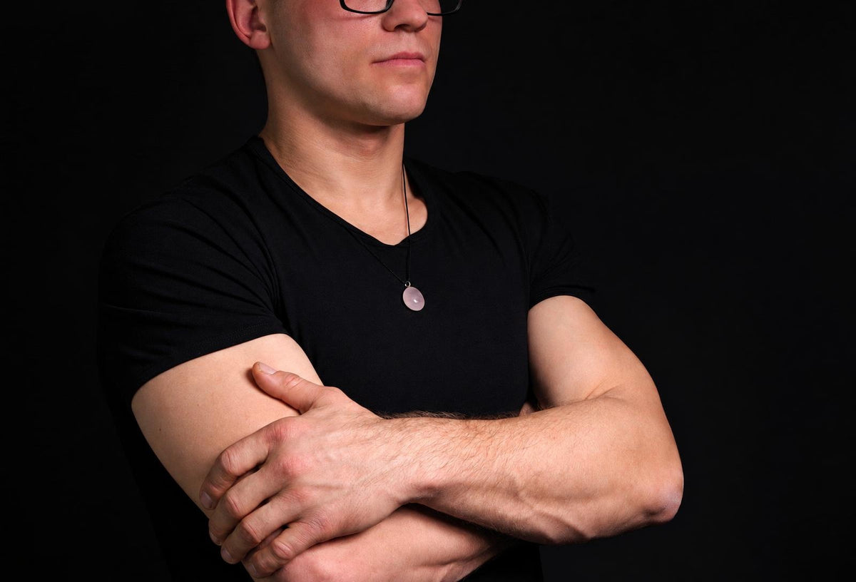 A man in a black t-shirt wears a necklace with a round, pale pink pendant on a dark surface.