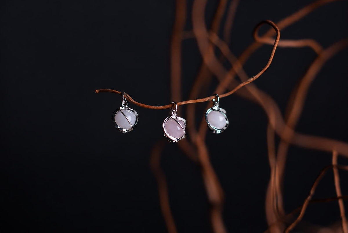 Three silver-toned pendants with pale pink stones hang from a curved brown branch against a dark background.