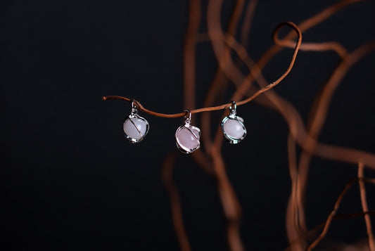 Three silver-toned pendants with pale pink stones hang from a curved brown branch against a dark background.