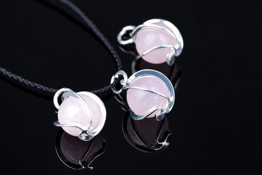 Three silver-toned wire pendants with pale pink spheres on black cords, arranged on a dark reflective surface.