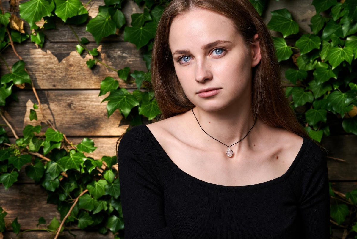 Woman wearing a black top and a necklace with a small round pale pink pendant against a wooden wall with green ivy.