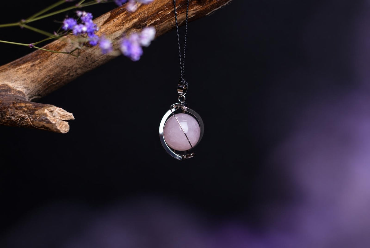 A silver-toned pendant with a pink sphere hangs from a dark cord on a wooden branch against a dark background.