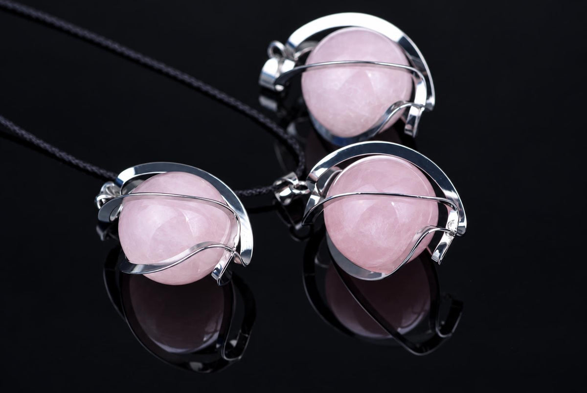 Three silver-toned pendants with pink spherical stones on dark cords, arranged on a reflective black surface.