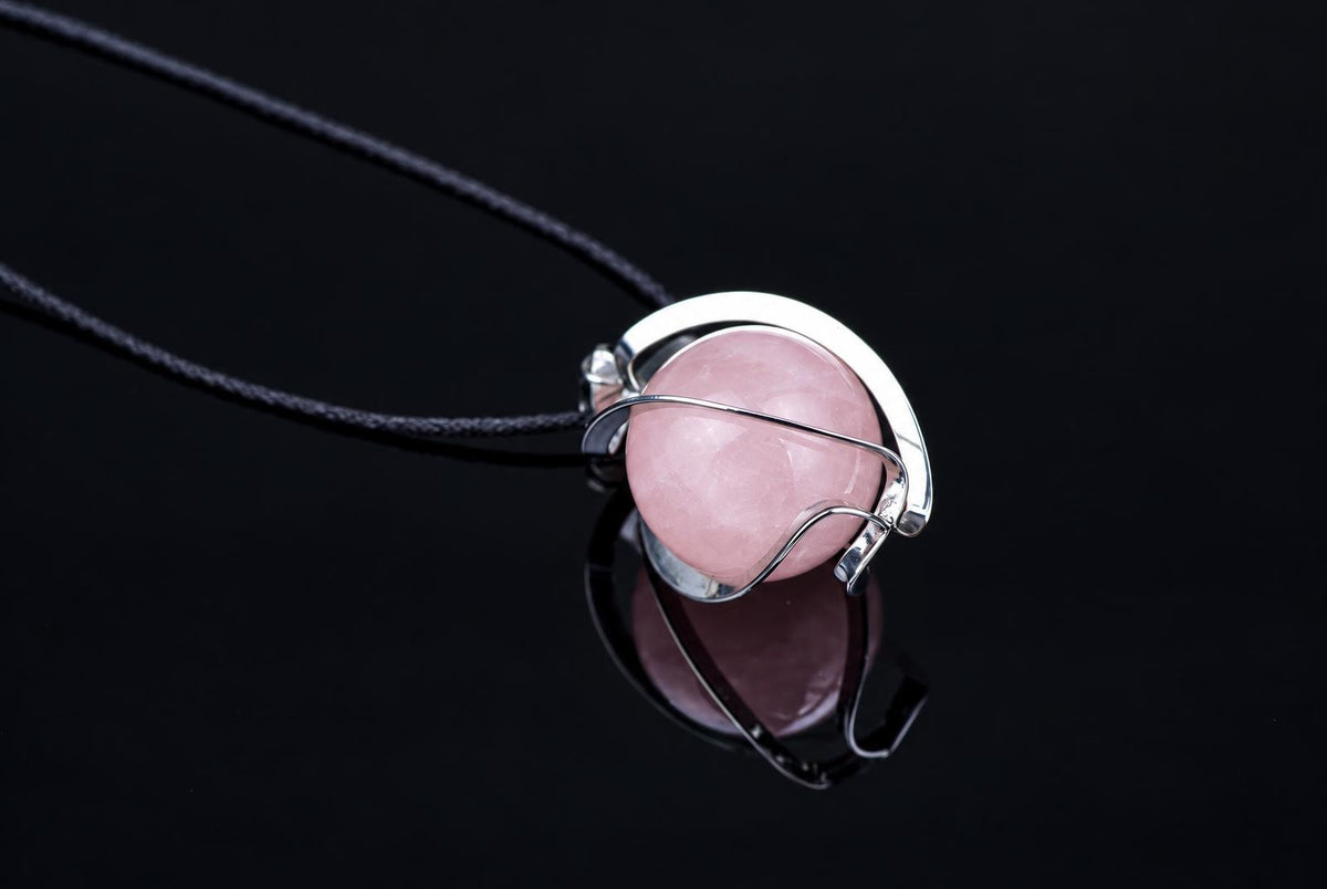 A silver-toned wire pendant holds a smooth, round pink stone on a black cord against a dark reflective surface.