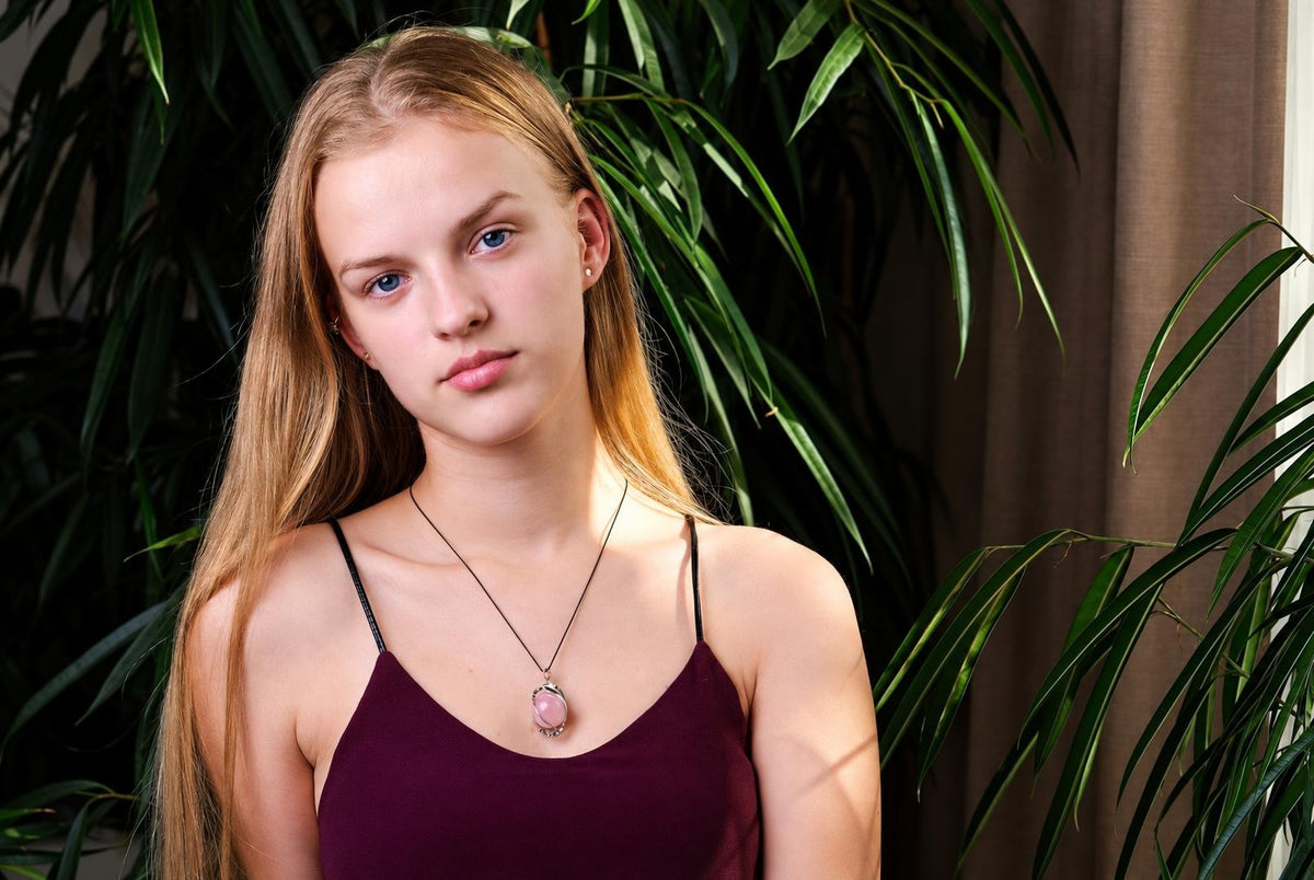 A woman wears a necklace with a pink oval pendant on a black cord, set against green plants and a dark surface.
