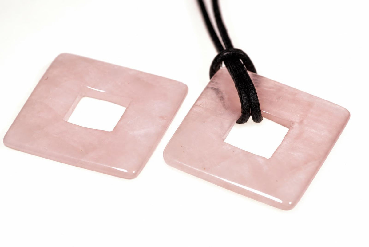 Two square, pink rose quartz pendants with central holes; one has a black cord attached. They rest on a white surface.