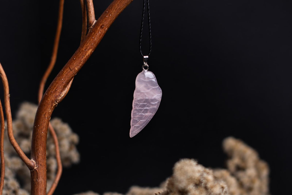 A pale pink carved pendant shaped like a wing hangs from a black cord on a dark surface with blurred branches.