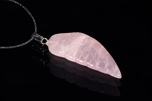 A pale pink, translucent pendant shaped like a wing hangs from a silver-toned chain on a dark surface.