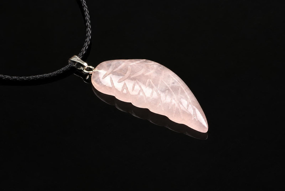 A pale pink carved pendant shaped like a wing, with engraved lines, hangs from a black cord on a dark surface.
