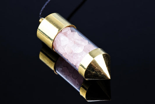 A clear tube pendant with pink rose quartz crystals, capped with gold-toned metal ends and a dark cord against a black surface.