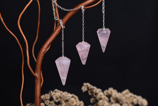 Three pale pink pointed stone pendulums hang from metal chains draped over a branch against a dark background.