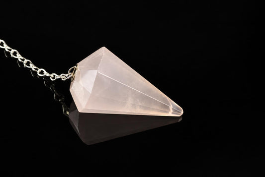 A faceted, pale pink crystal pendant on a silver-toned chain rests on a dark, reflective surface.