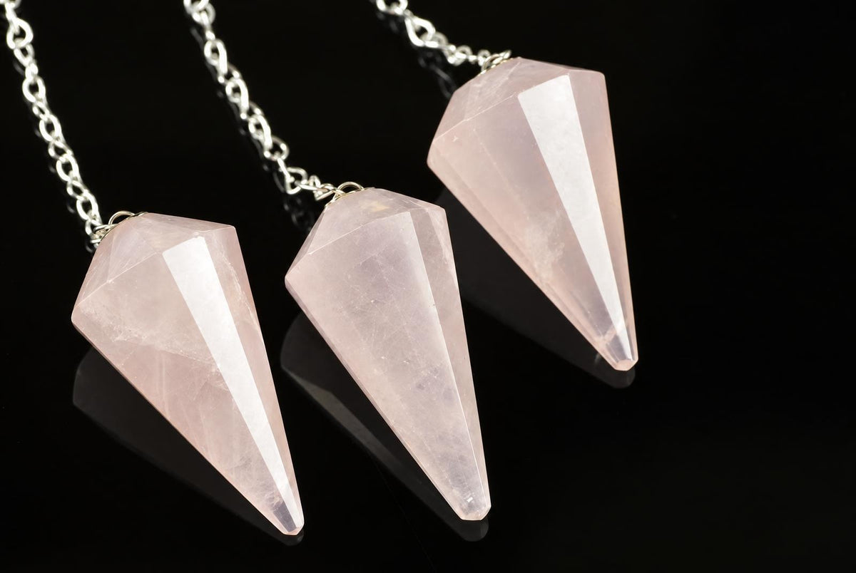 Three faceted, pale pink rose quartz pendulums on silver-toned chains rest on a dark surface.