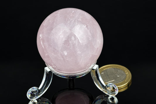 A pink rose quartz sphere rests on a clear glass stand against a dark background, with a coin nearby for scale.