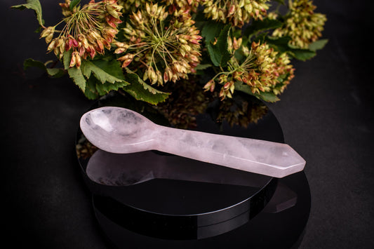 A pale pink, translucent spoon carved from stone rests on a dark surface with greenery in the background.