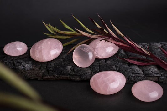 Six polished pink rose quartz stones of varying oval and round shapes rest on a dark wooden surface with greenery.