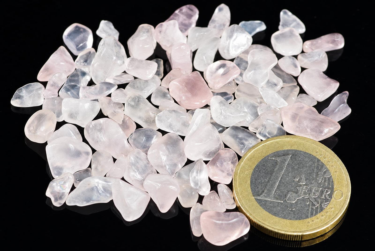 A pile of small, translucent pink and white rose quartz tumbled stones on a dark surface next to a one euro coin.