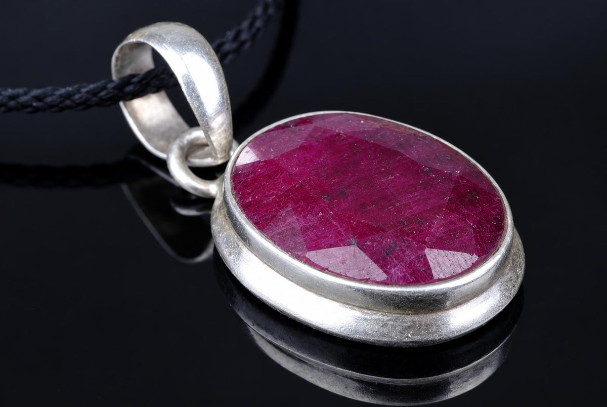 A silver-toned pendant with a large oval red stone on a black cord, resting on a dark reflective surface.