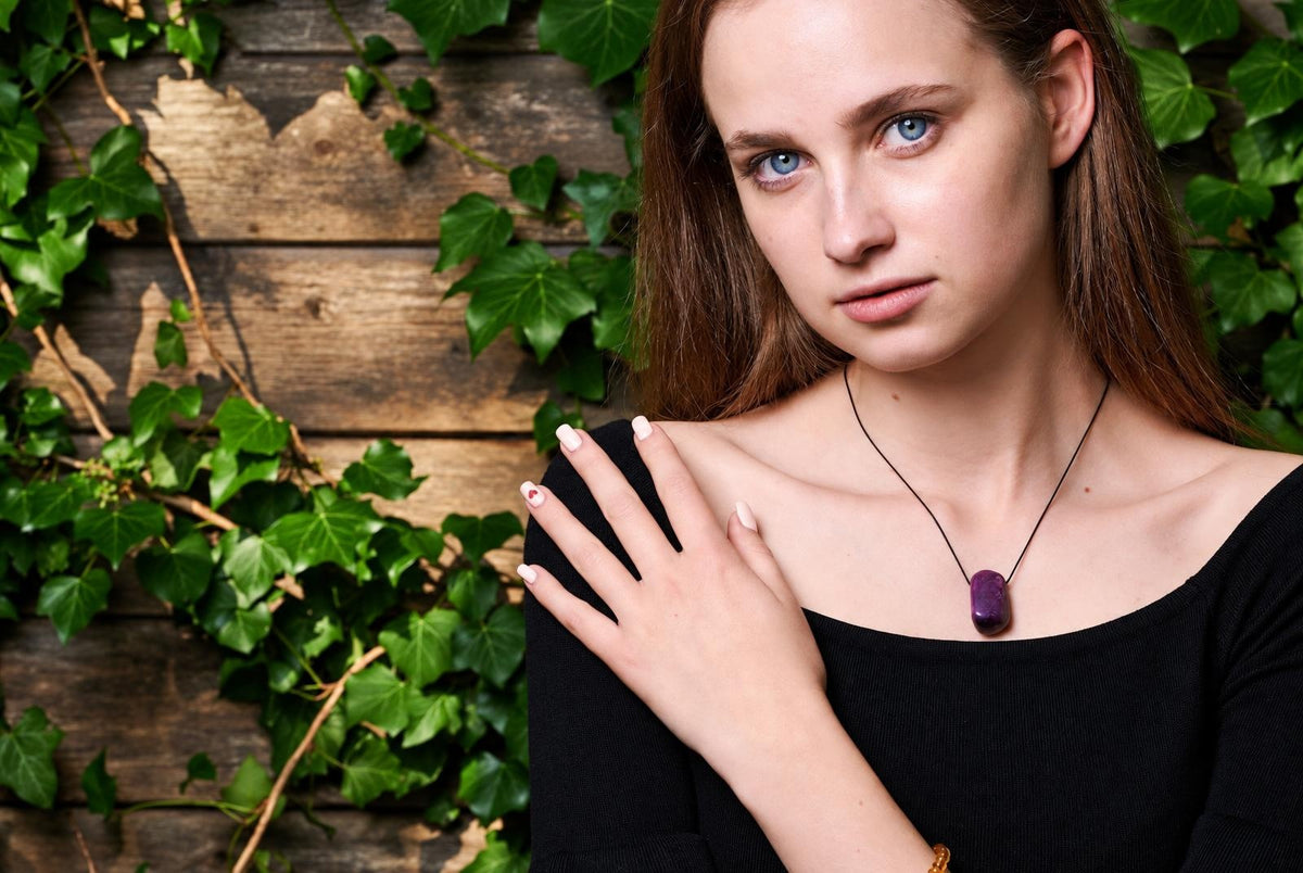 A woman wears a black top and a dark purple pendant on a thin cord, set against a wooden surface with green ivy.